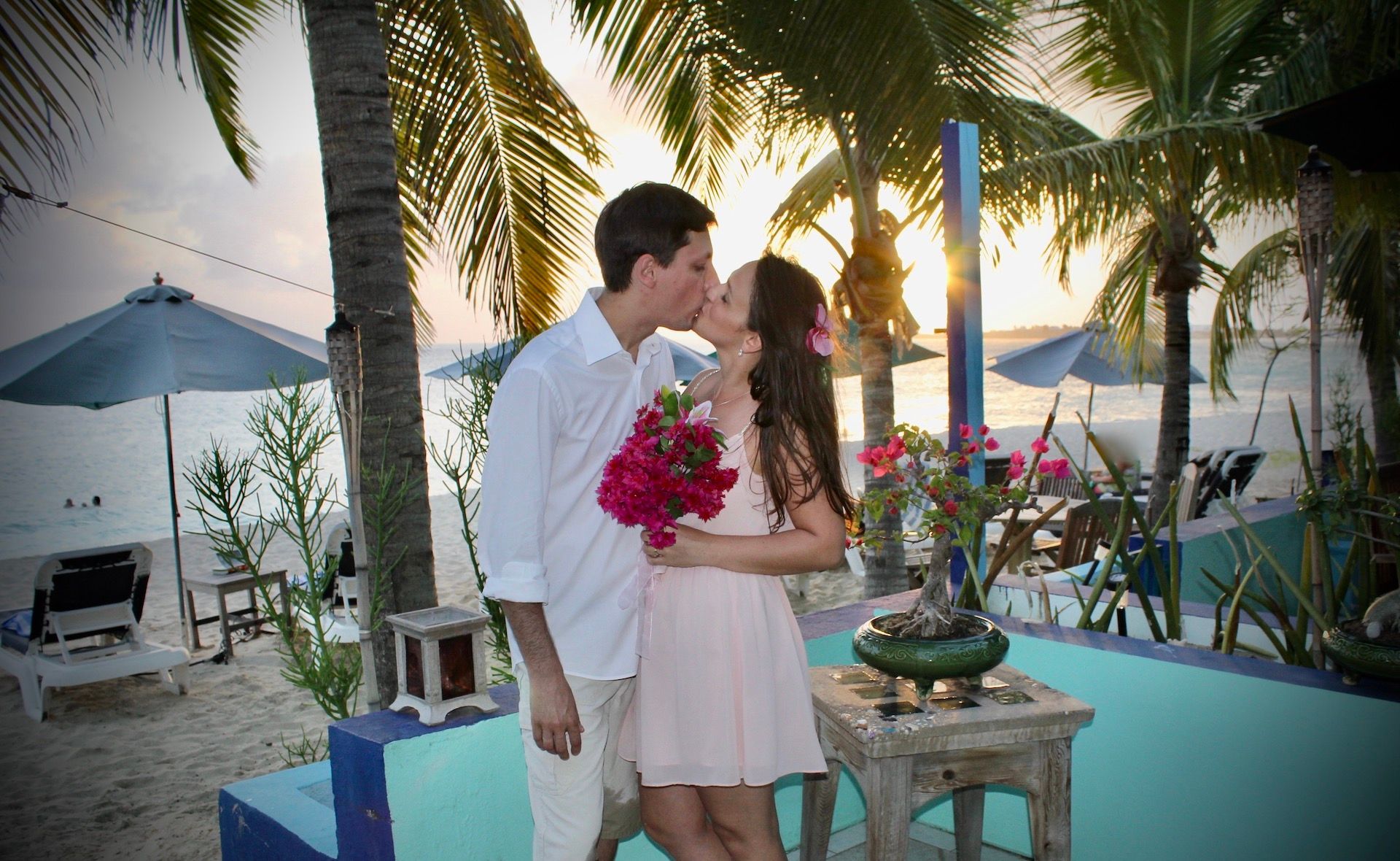 Couple kissing on a beach at sunset, holding flowers. Palm trees and blue pool in background.