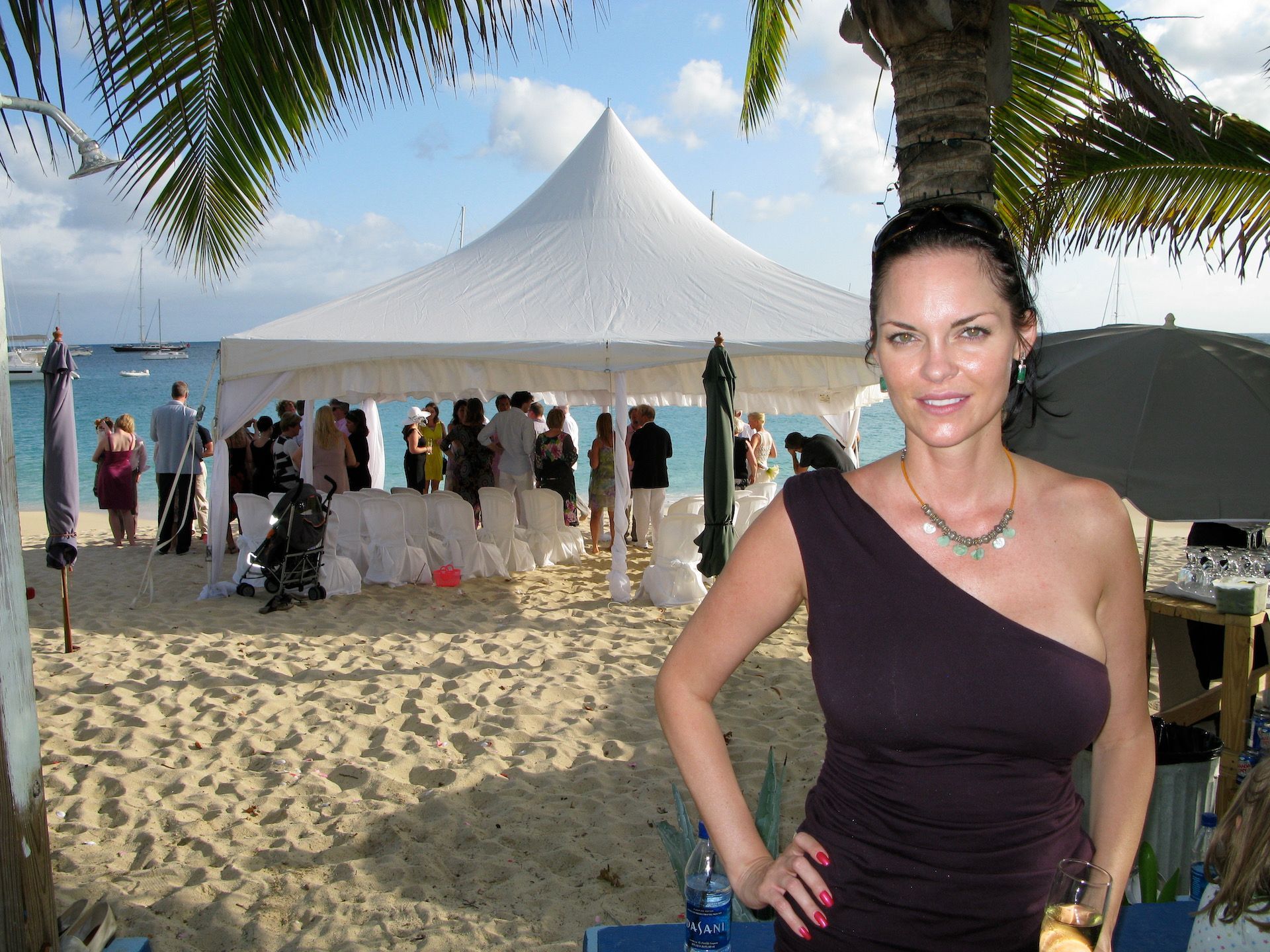 Woman in purple dress on a beach with wedding tent and guests in the background.