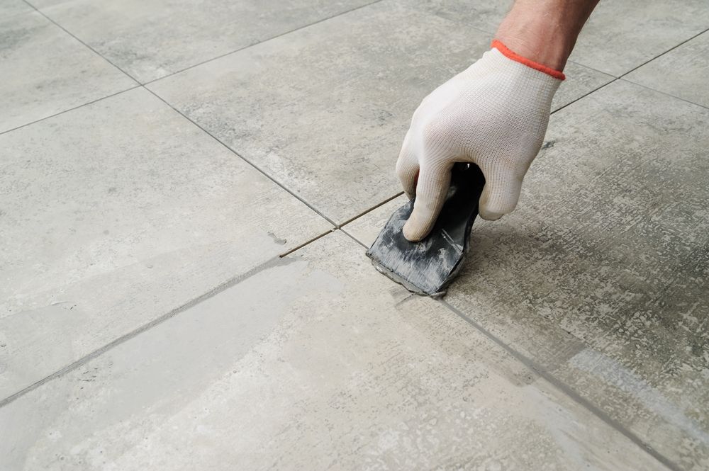 Gloved Hand Using a Trowel to Apply Grout Between Gray Floor Tiles — Tropic Tiling in Malanda, QLD
