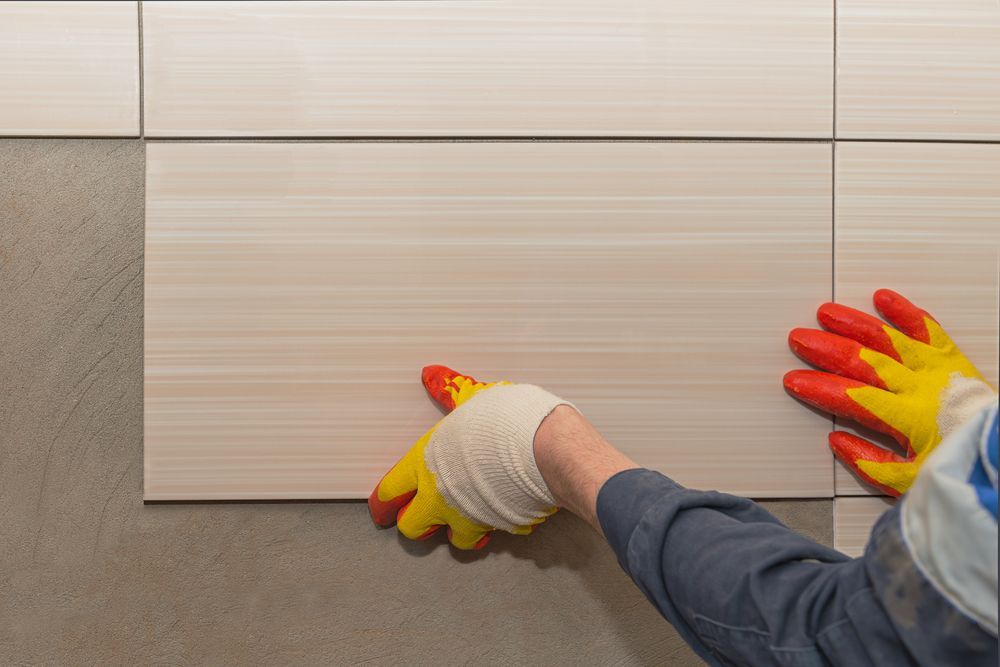 Person With Gloved Hands Installing Beige Tile on a Gray Wall — Tropic Tiling in Tolga, QLD