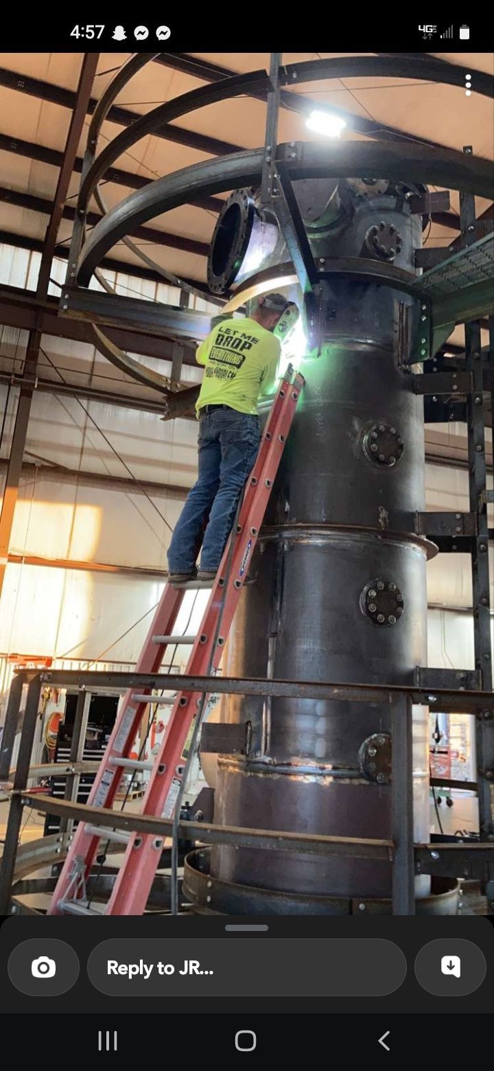 A man is welding a large pipe in a factory while standing on a ladder.