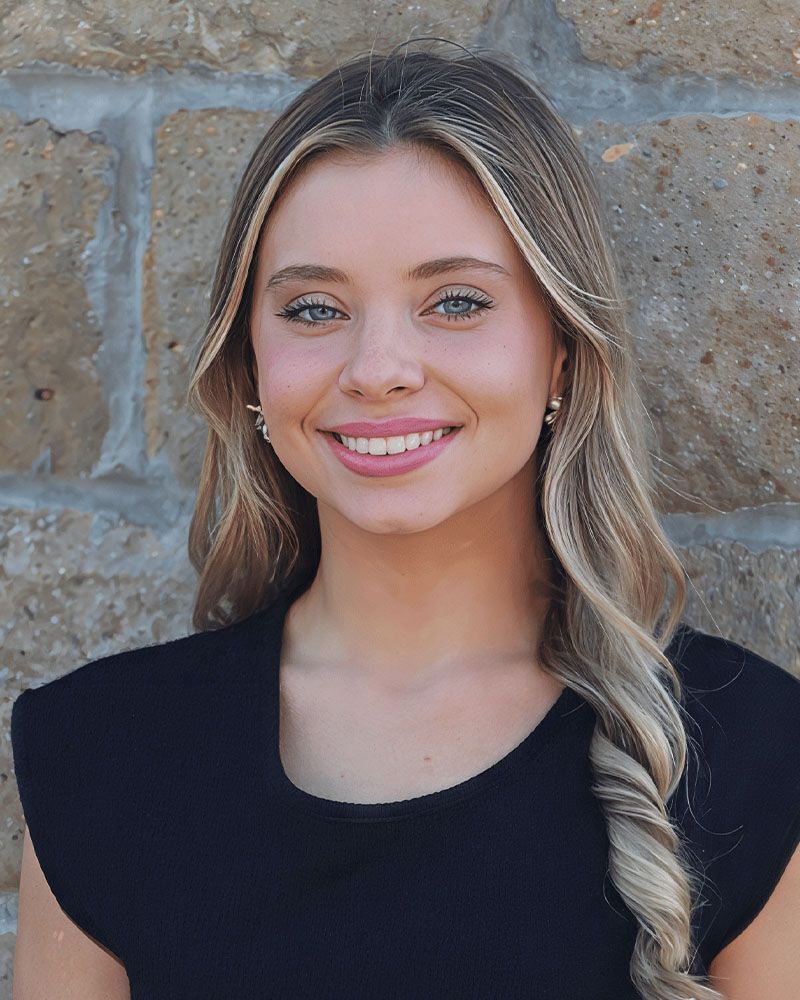 A woman wearing a one shoulder top is smiling in front of a brick wall.