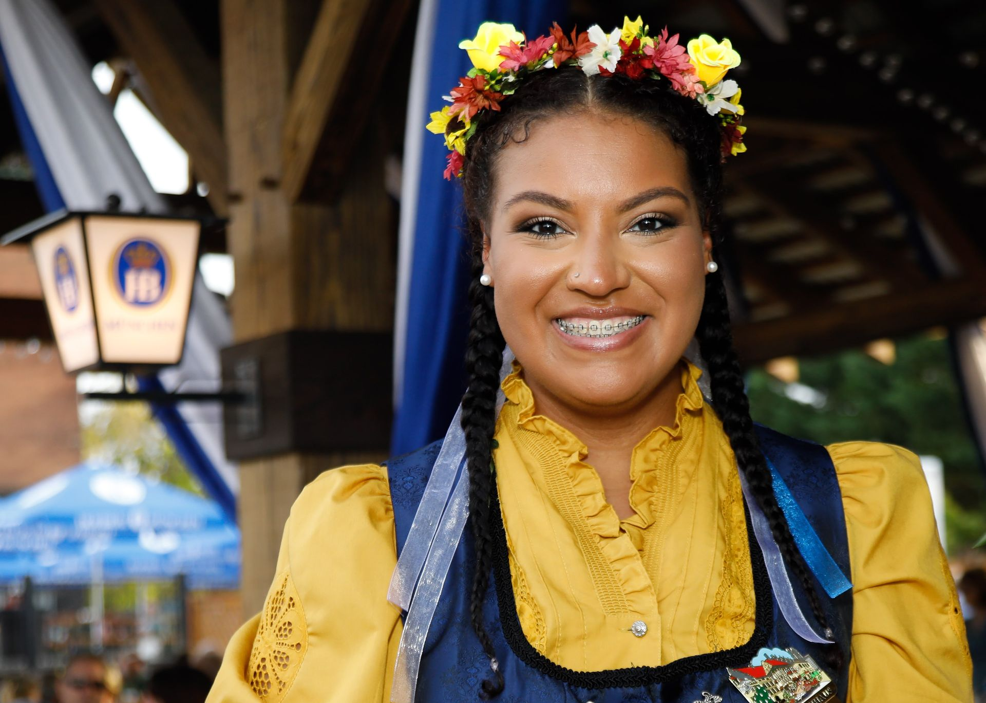 Woman in yellow and blue traditional outfit, floral crown, and braces smiles outdoors at a festival.