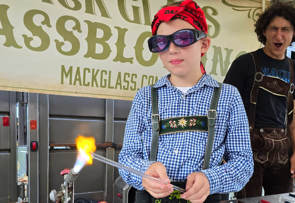 Boy in safety glasses and bandana doing glassblowing, adult watches. Outdoors.