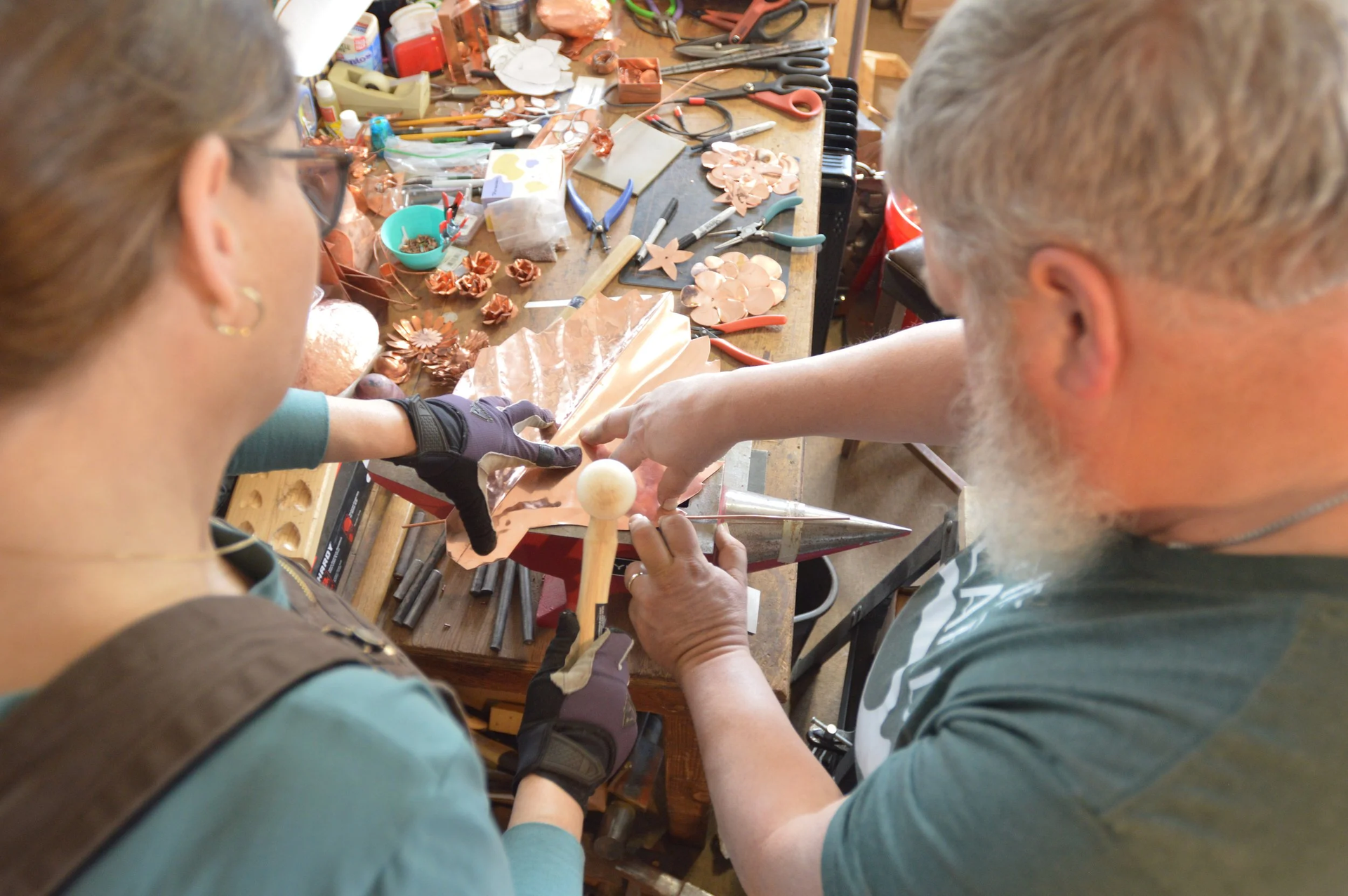 Two people work on a blacksmithing project at a cluttered workbench. One holds the piece, the other works with it.