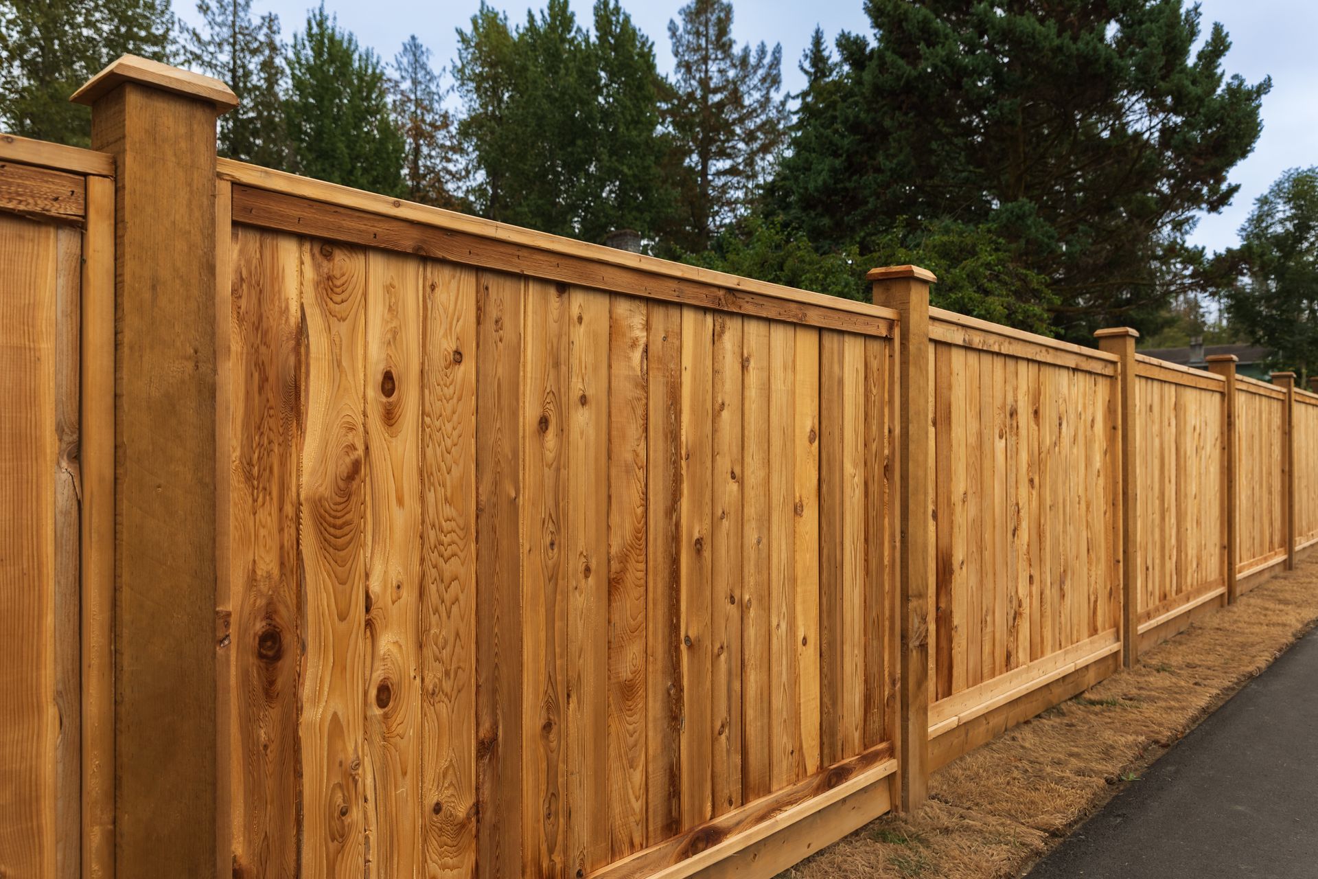 A wooden fence is sitting next to a road with trees in the background.