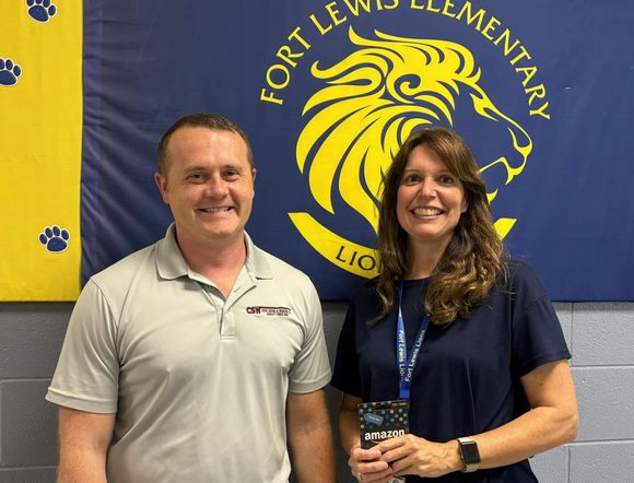 A man and a woman are standing in front of a flag that says fort lewis elementary