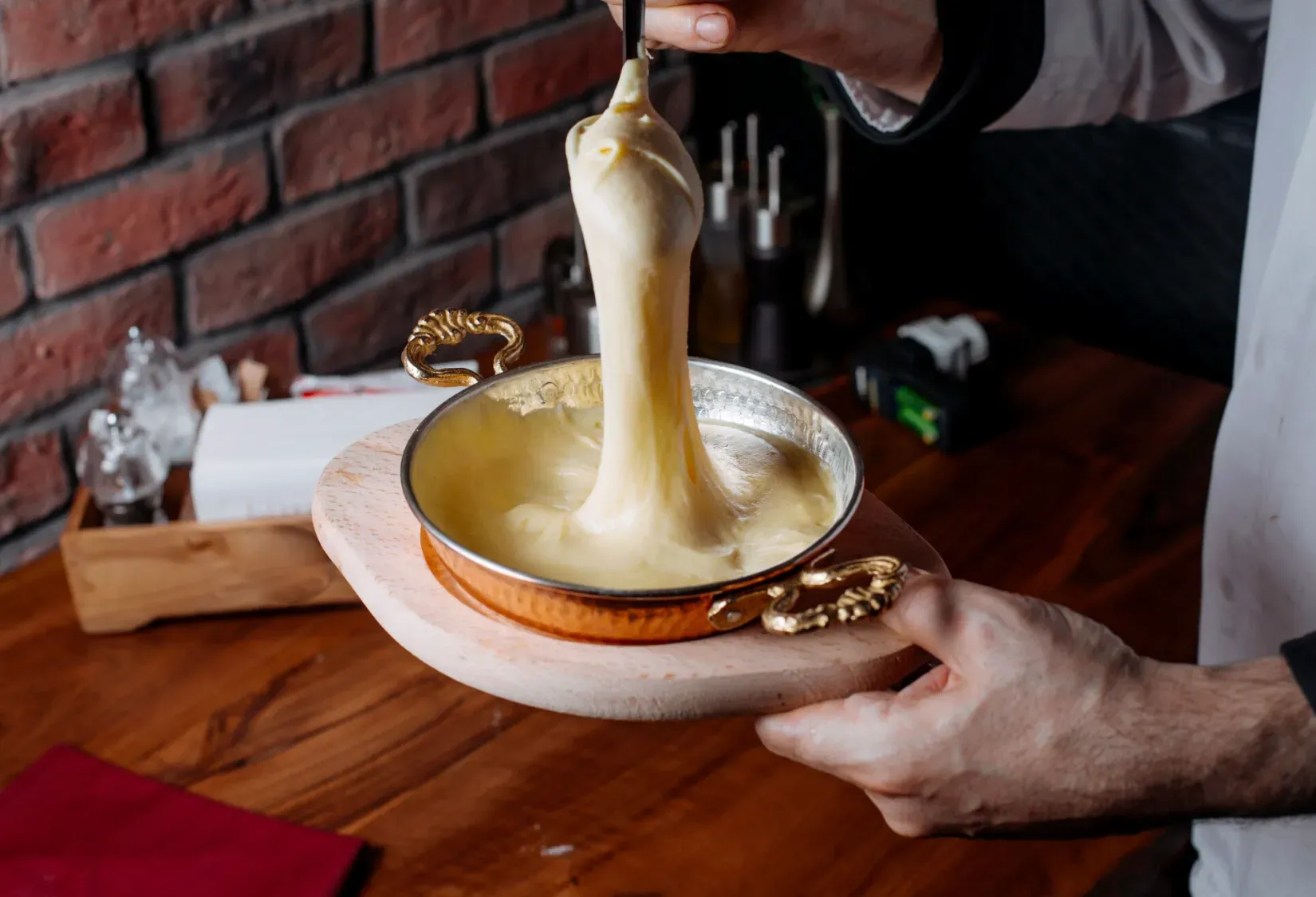 Mão de um chef segurando um recipiente de metal onde o queijo derretido do fondue é puxado para cima
