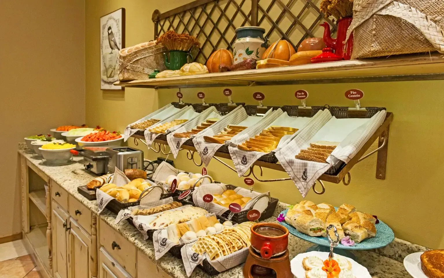 Buffet de café da manhã com diversos tipos de pães, doces e acompanhamentos em um balcão com prateleira suspensa.