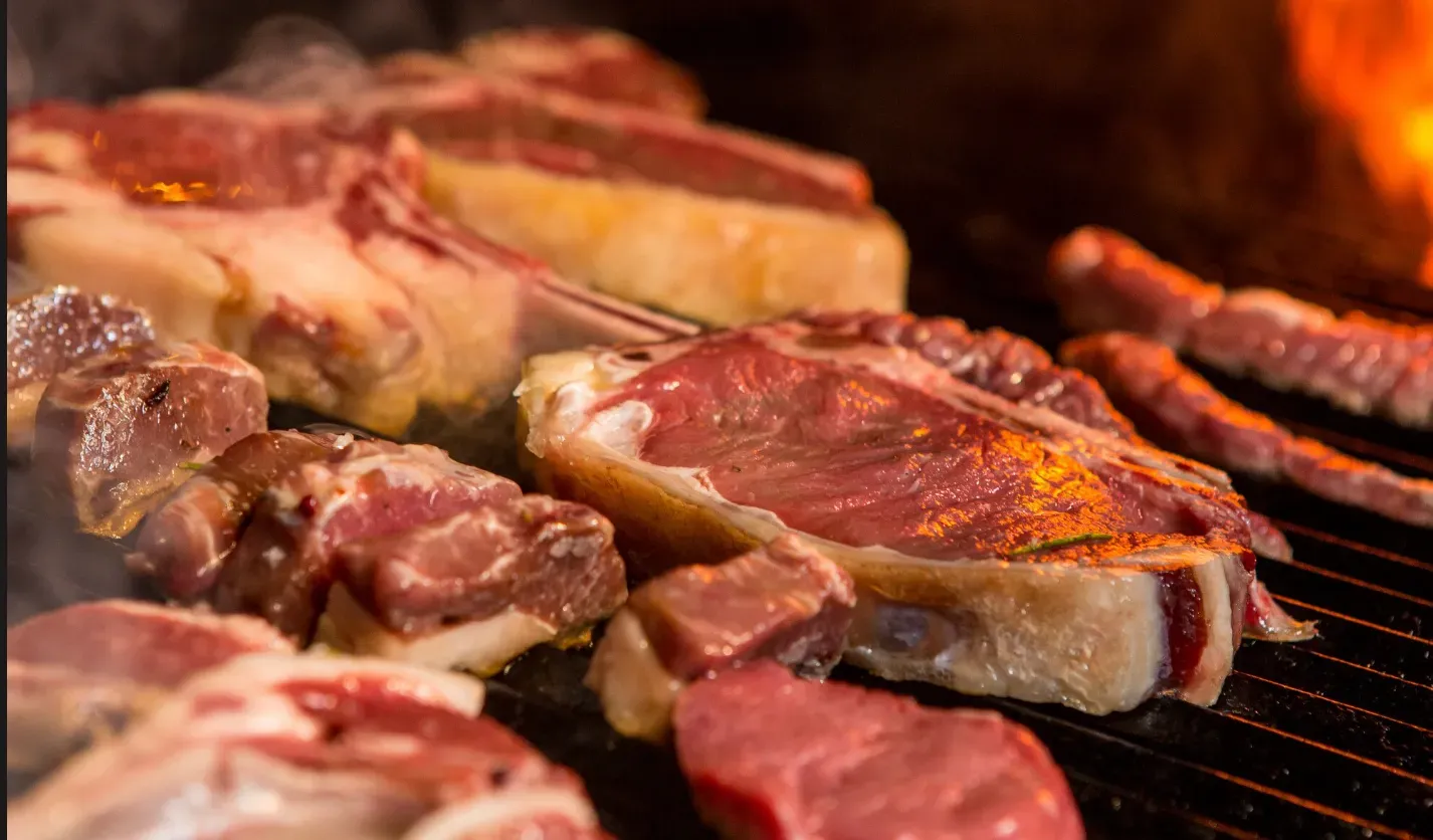 Cortes nobres de carne vermelha e linguiças sendo assados na grelha sobre chamas vivas, representand