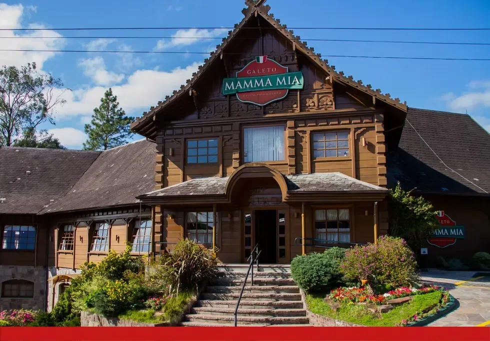 Fachada diurna do restaurante Galeto Mamma Mia em Gramado. O edifício possui uma arquitetura clássica em estilo alpino europeu, construído em madeira escura com detalhes entalhados e telhado bem inclinado. Na frente, há uma escadaria de pedra cercada por jardins bem cuidados com flores coloridas e arbustos. No topo, destaca-se o letreiro verde e vermelho com o nome do local.