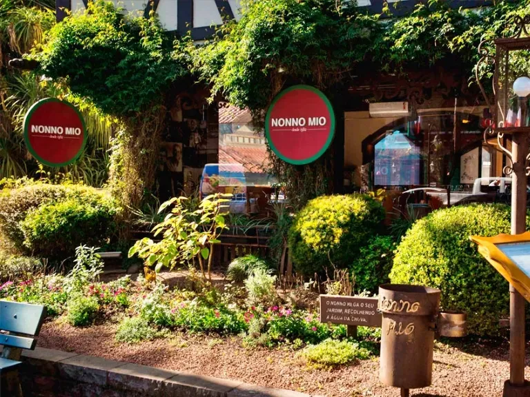 Fachada externa do restaurante Nonno Mio durante o dia. A entrada é cercada por uma densa vegetação verdejante, com arbustos podados em formato arredondado, plantas trepadeiras cobrindo a estrutura e um pequeno jardim com flores. Duas placas redondas com as cores da bandeira da Itália (verde, branco e vermelho) exibem o nome 