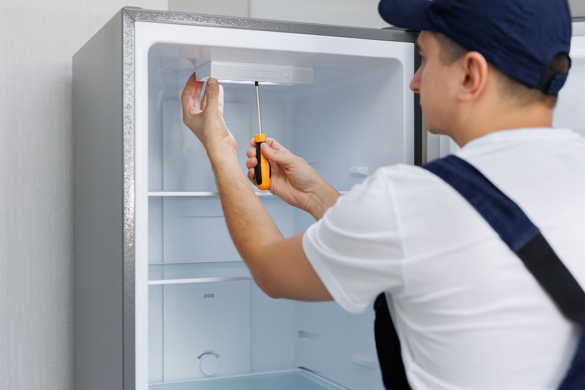 Man in a uniform repairs the light in the refrigerator with a screwdriver