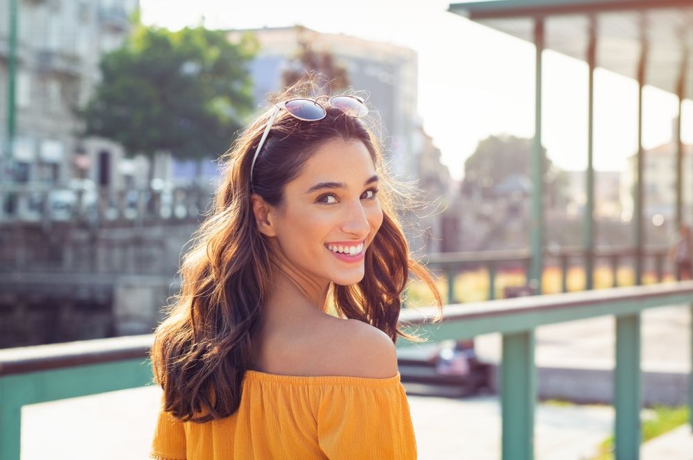 A woman in a yellow off the shoulder top is smiling and looking over her shoulder.