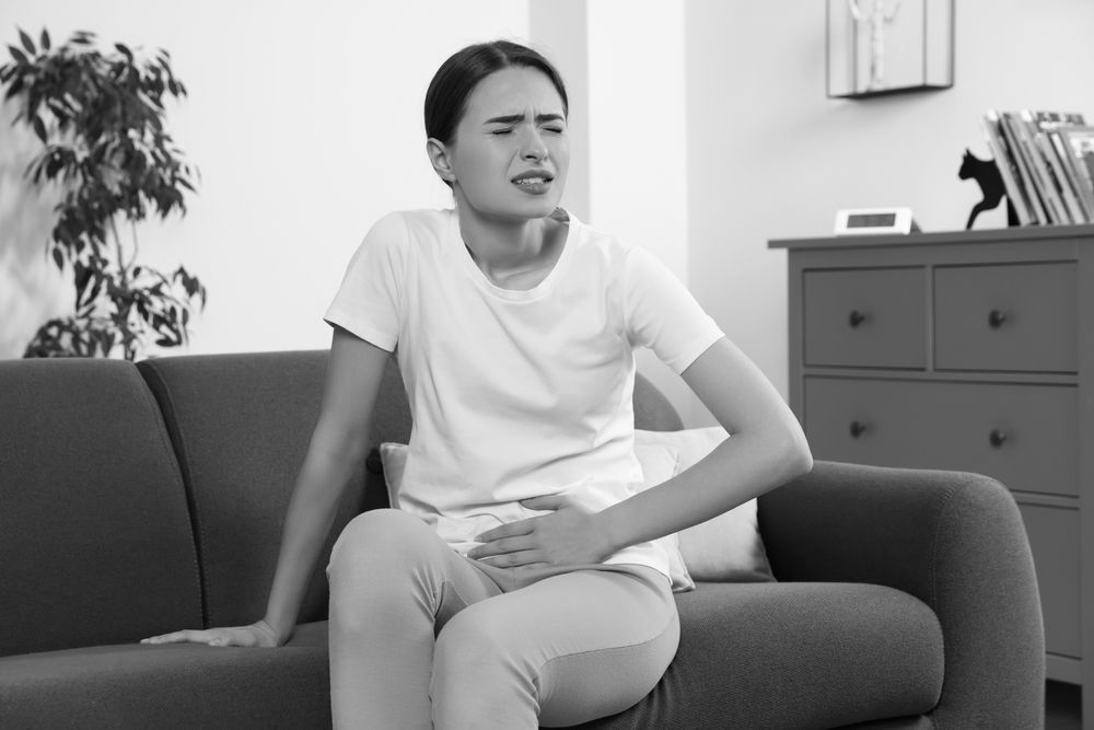 A woman is sitting on a couch holding her stomach in pain.