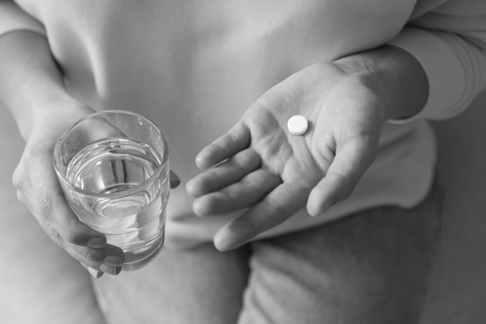 A person holds a glass of water in one hand and a single round white pill in the palm of their other hand.