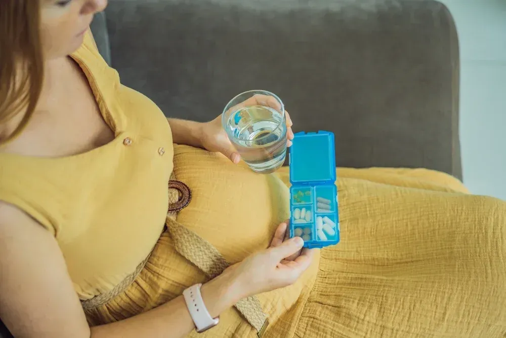 A pregnant woman is sitting on a couch holding a glass of water and a pill box.