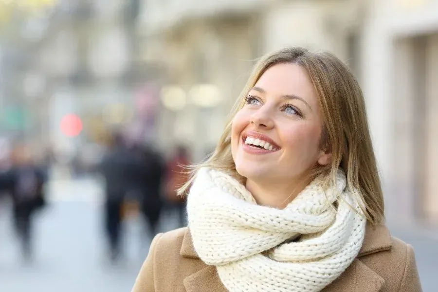 A woman wearing a scarf and a coat is smiling on a city street.