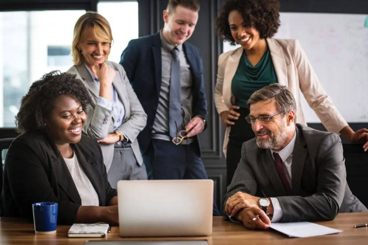 A professional team in business attire gathers around a laptop in an office, smiling as they collaborate on a project.