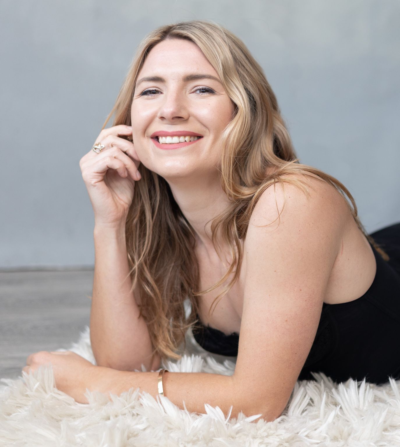A woman in a black dress is laying on a white rug and smiling.