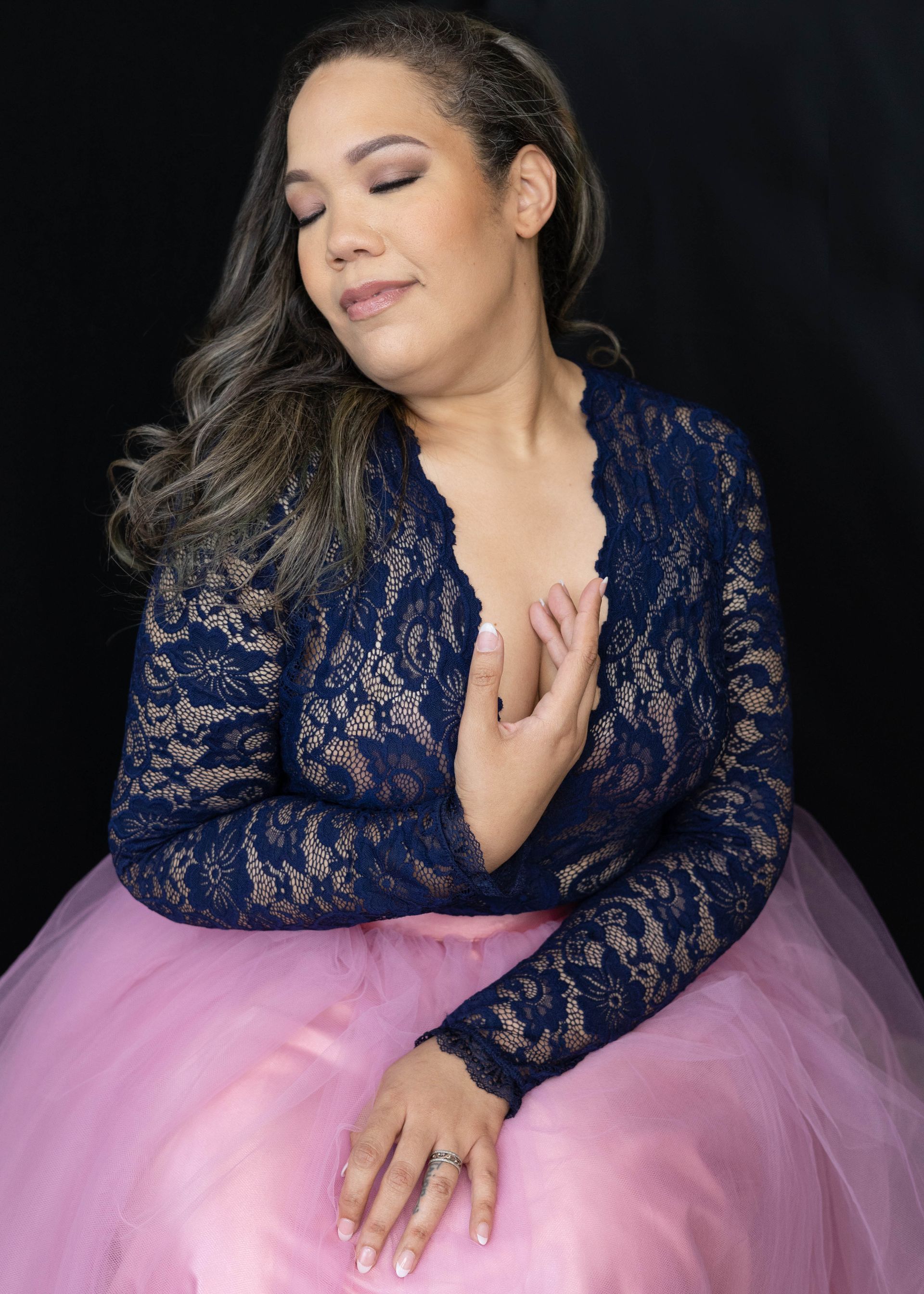 A woman in a blue lace top and pink skirt is sitting with her eyes closed.