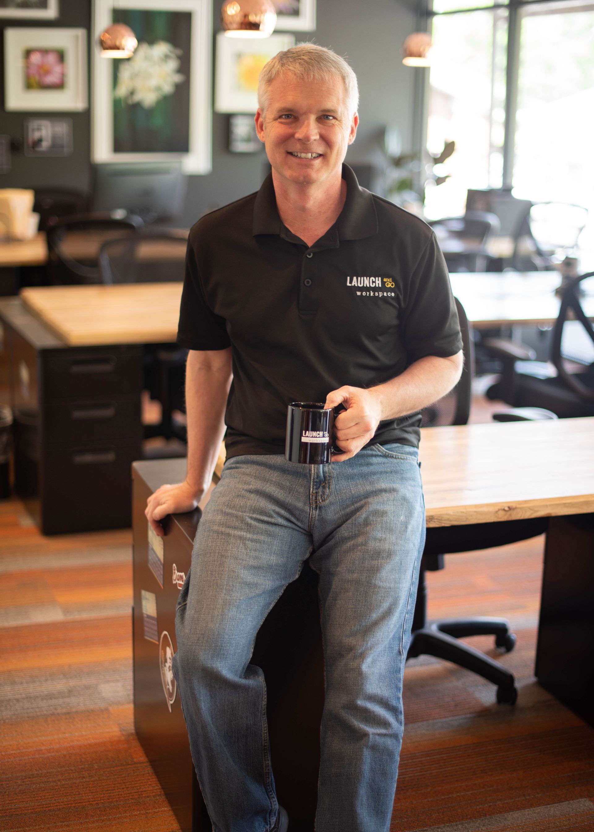A man is sitting on a desk in an office holding a cup of coffee.