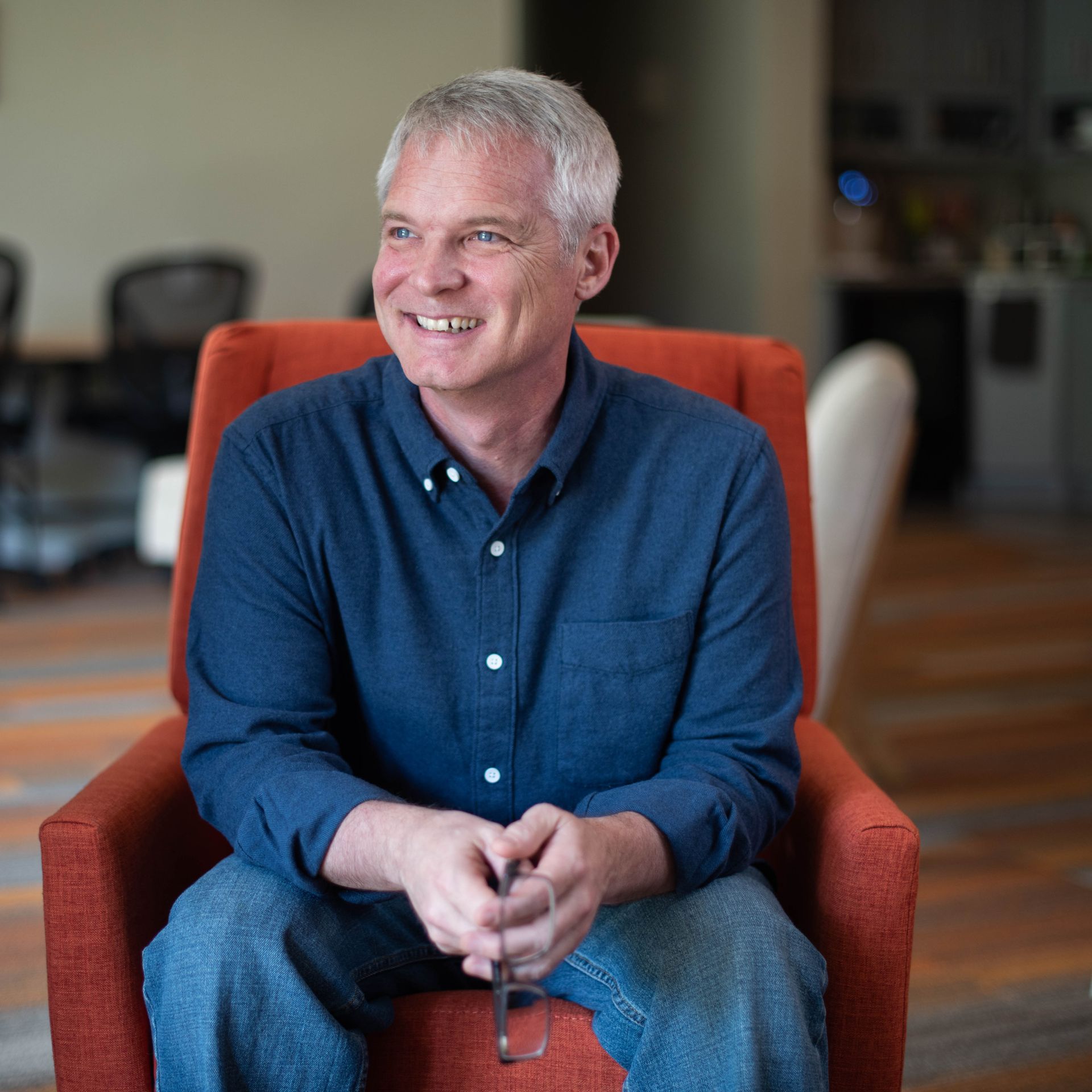 A man in a blue shirt is sitting in an orange chair and smiling.