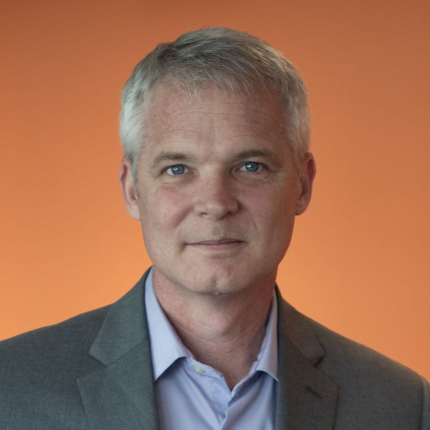 A man in a suit and a purple shirt is standing in front of an orange wall.