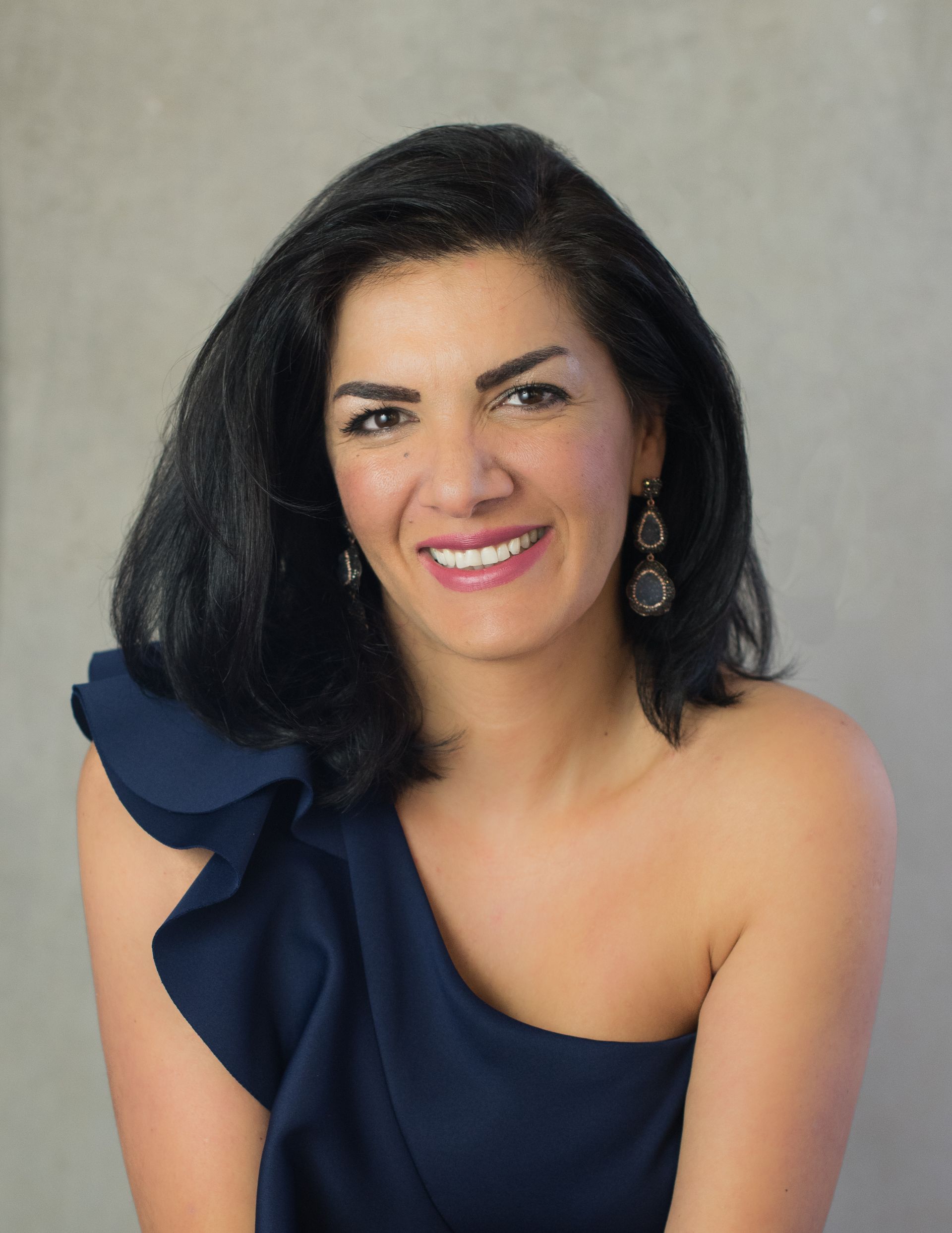 A woman wearing a blue one shoulder top and earrings smiles for the camera.