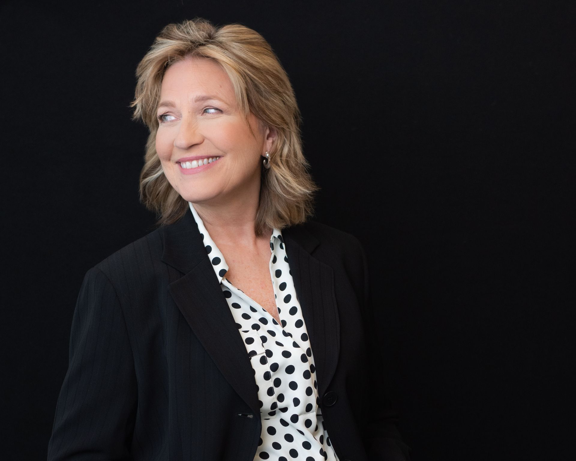 A woman in a black jacket and polka dot shirt is smiling.