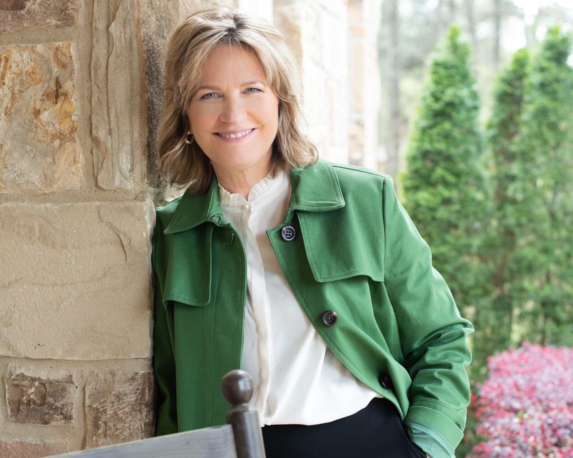 A woman in a green jacket is leaning against a stone wall and smiling.
