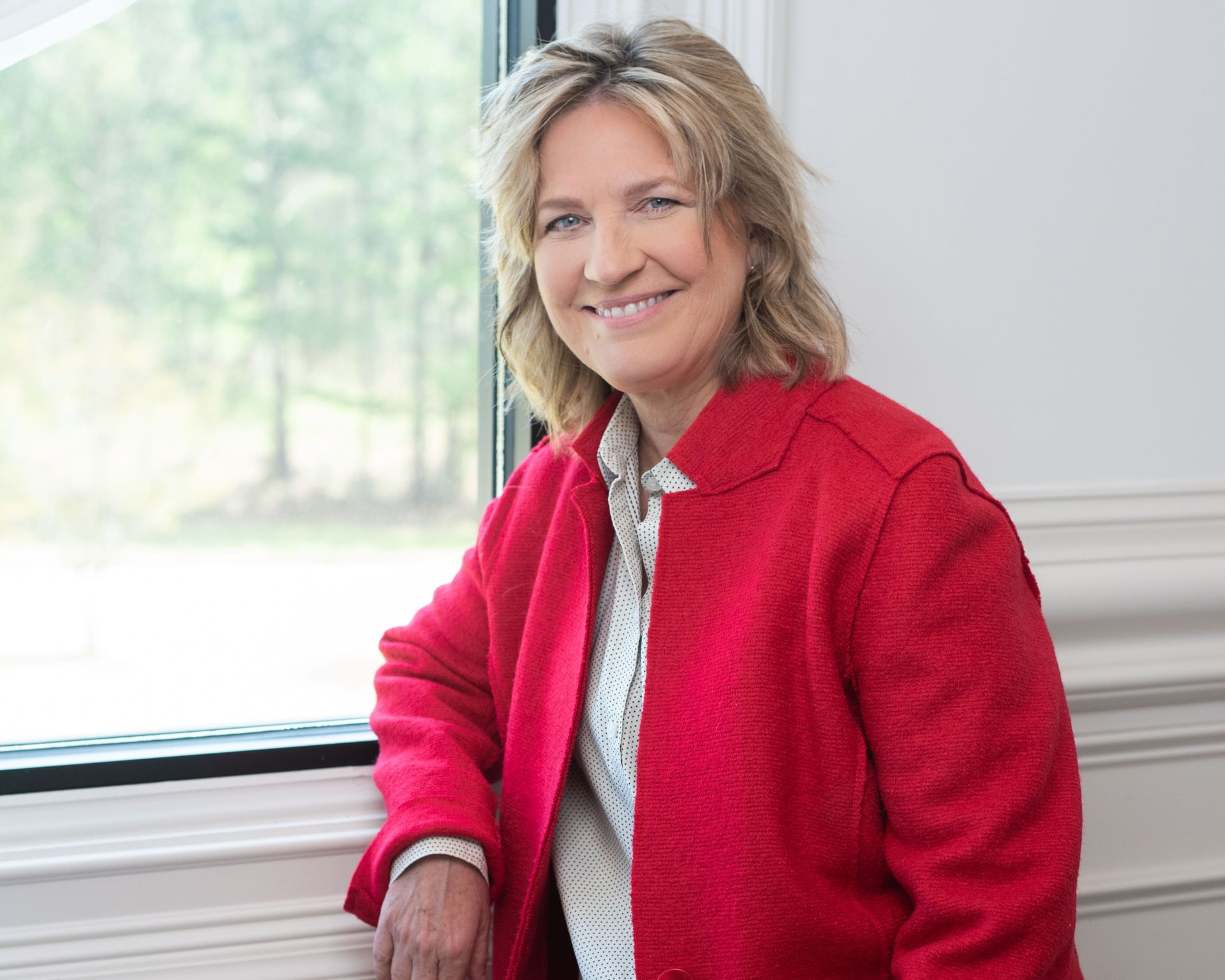 A woman in a red coat is leaning against a wall and smiling.