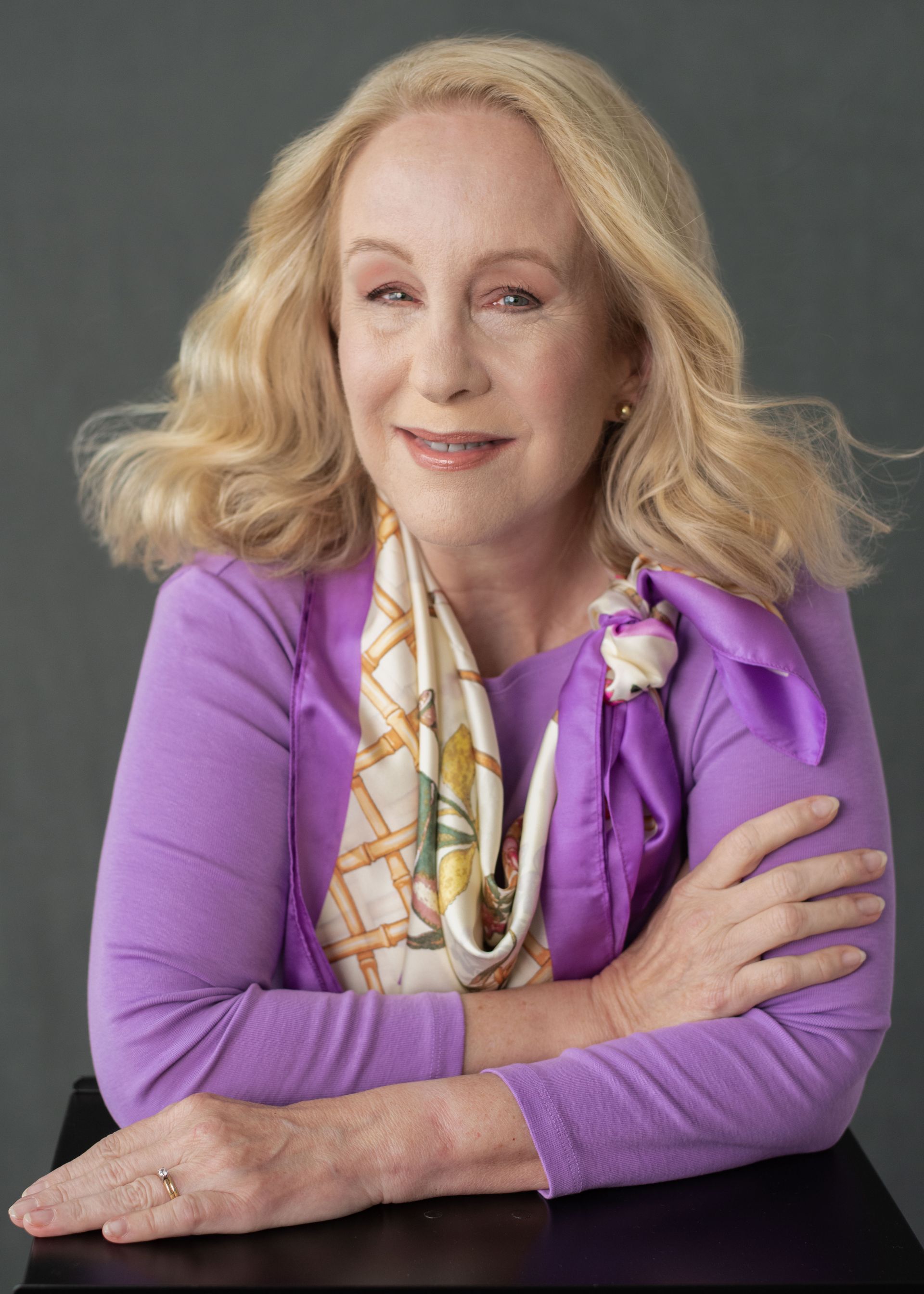A woman in a purple sweater and scarf is sitting at a table with her arms crossed.