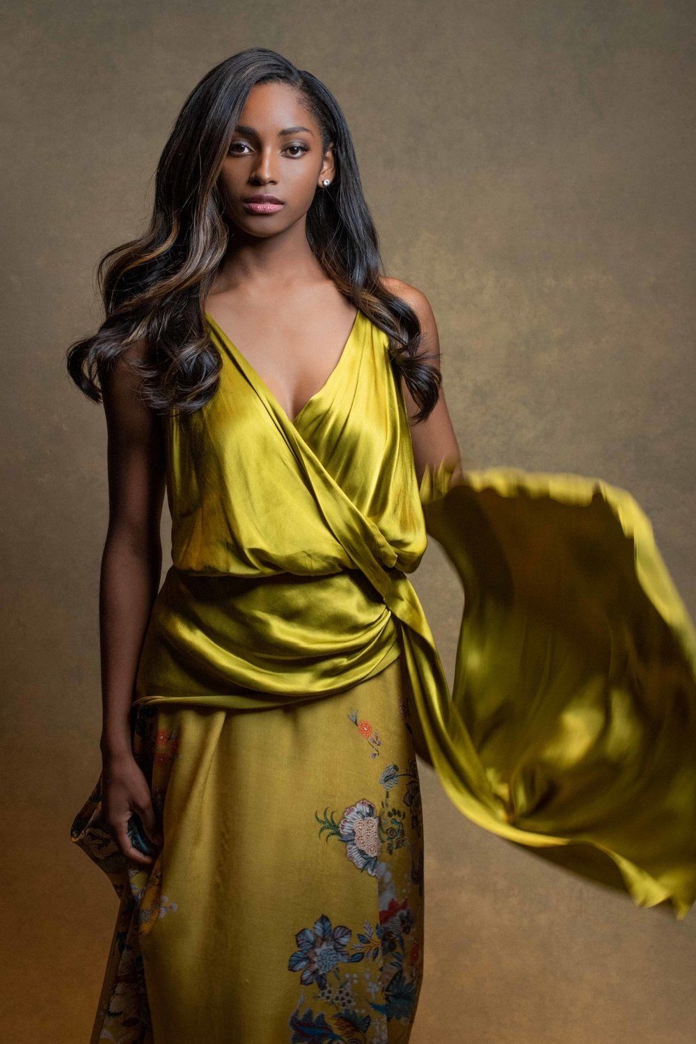 A woman in a yellow dress is standing in front of a brown background.