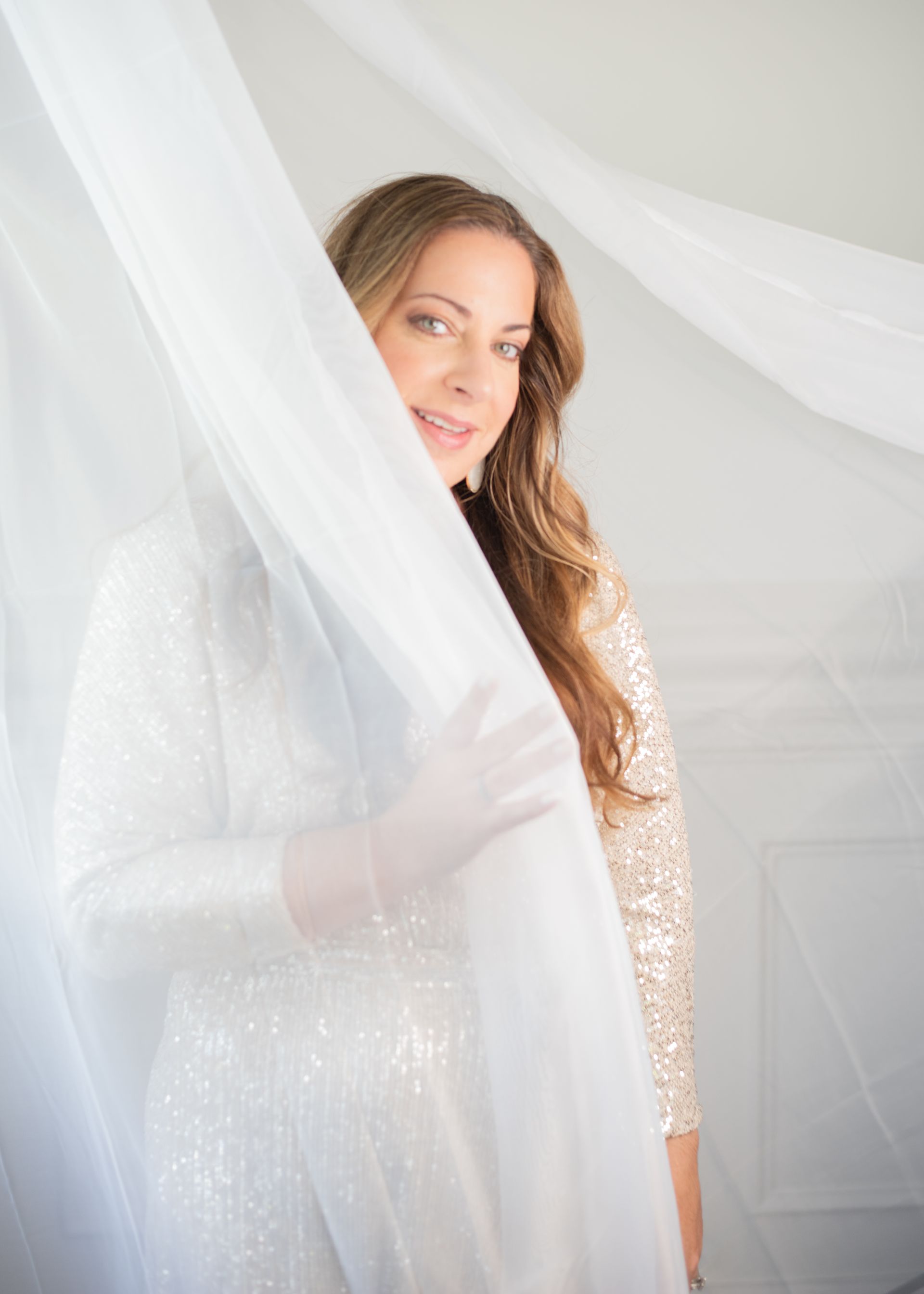 A woman in a sequined dress is standing behind a white curtain.