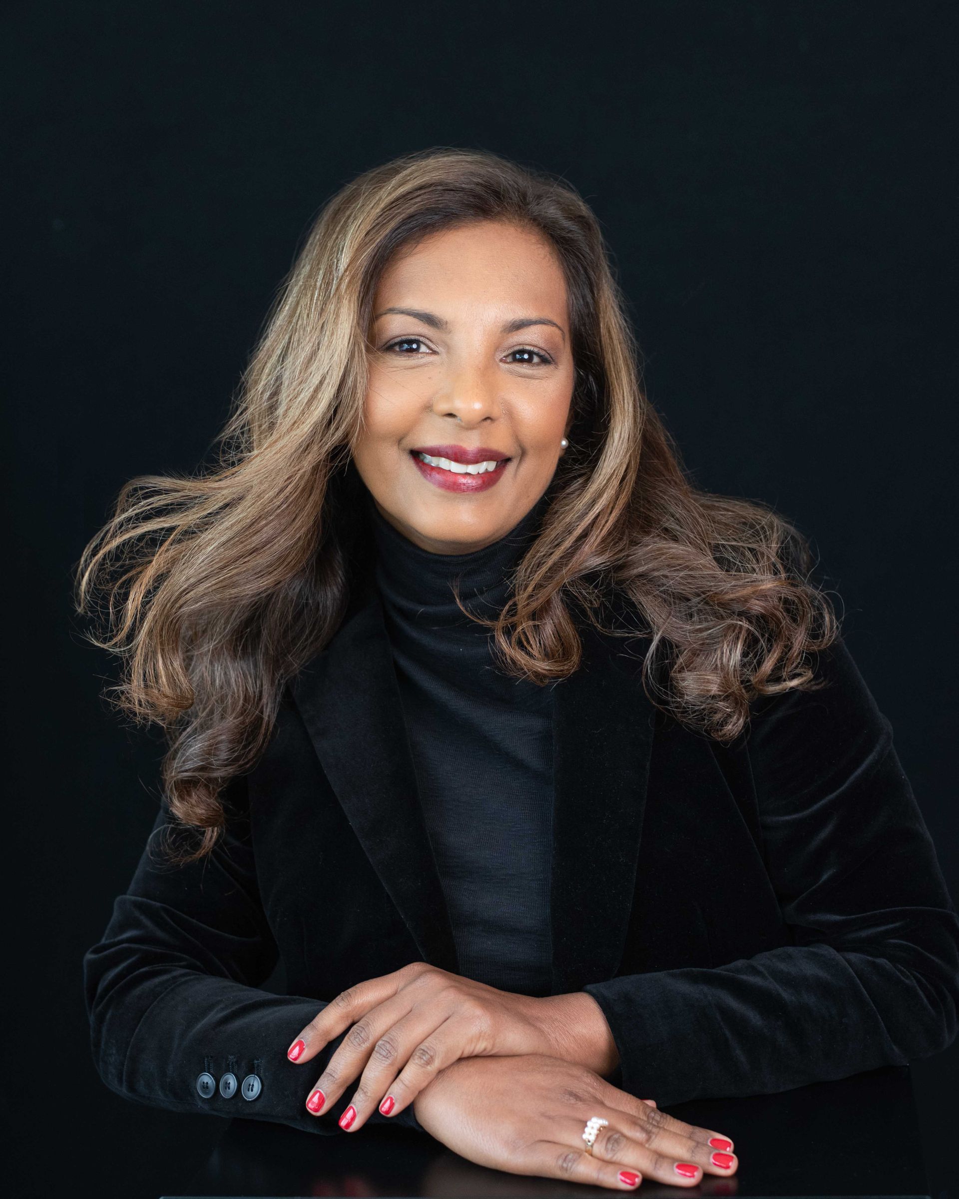 A woman in a black jacket and turtleneck is sitting at a table with her hands folded.