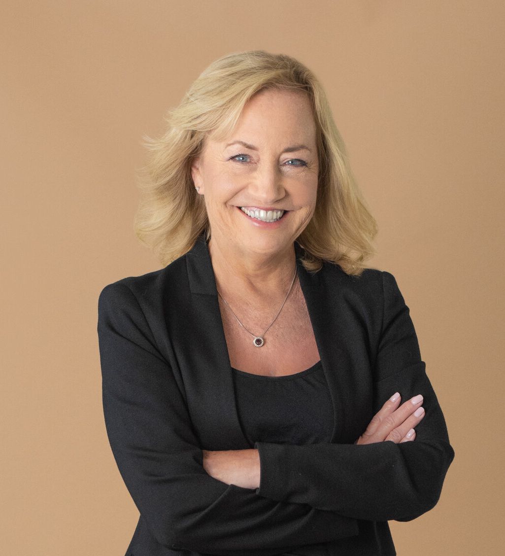 A woman in a black jacket and necklace is smiling with her arms crossed.