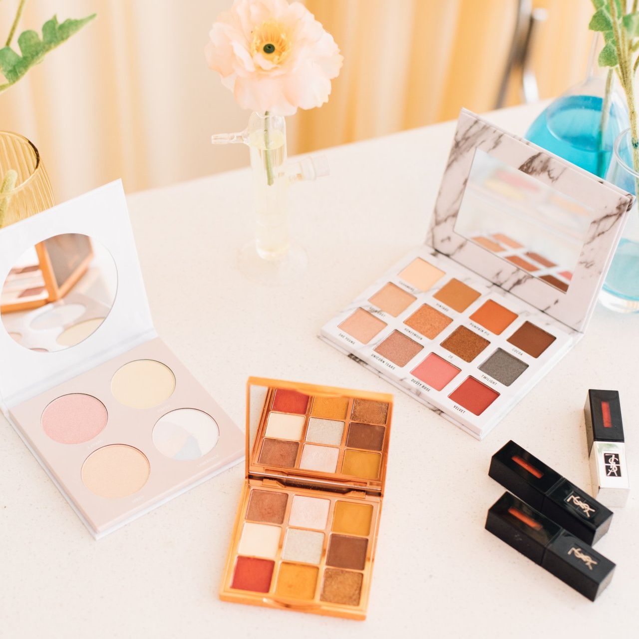 Several makeup palettes are on a table with a vase of flowers in the background