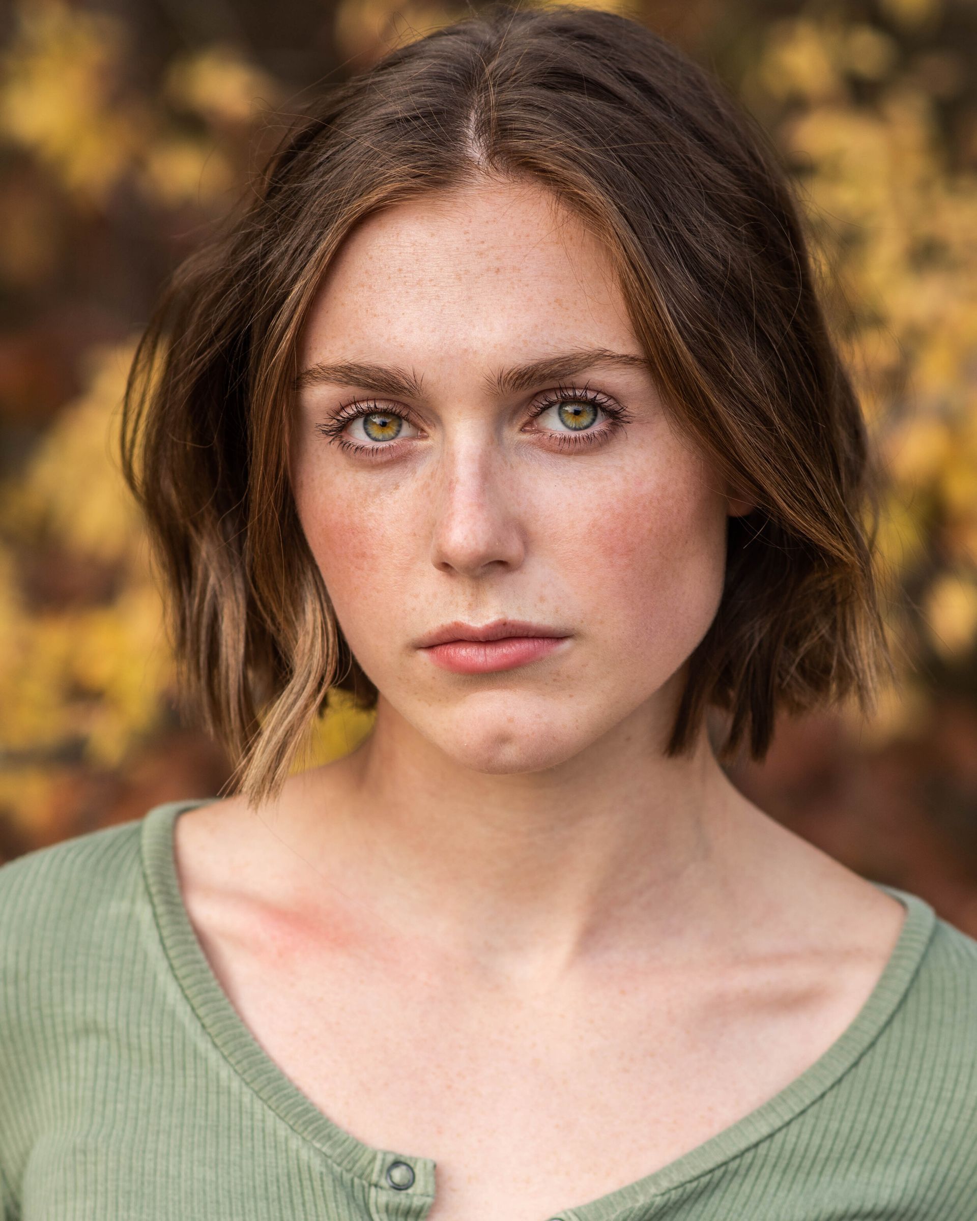 Woman with short brown hair, wearing a green shirt, looking directly at the camera with a neutral expression. Blurred background.