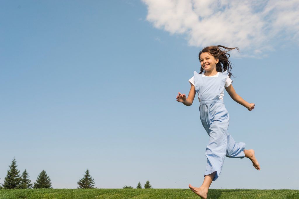 Girl with curly hair running barefoot on green grass under a blue sky with a cloud.
