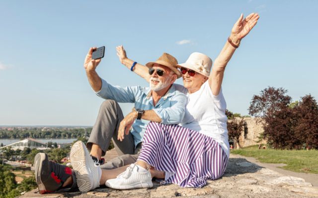 Elderly couple takes a selfie outdoors, arms raised, smiling. One wears a hat, sunglasses. Blue sky background.