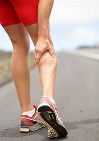 Runner gripping calf muscle, likely in pain, on a road. Red shorts, running shoes.