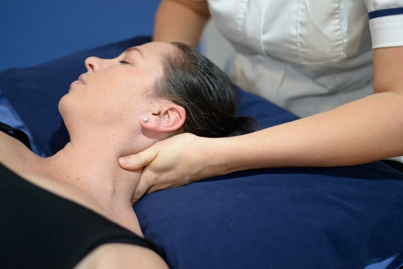 Woman receiving neck massage; therapist's hands on patient's neck. Setting: blue pillow, white shirt.