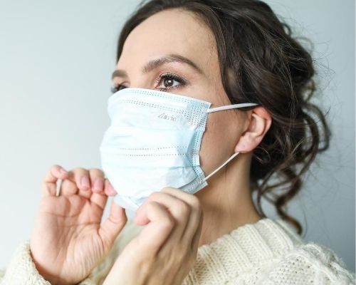 Woman adjusting a blue surgical mask, wearing a white sweater, indoors.
