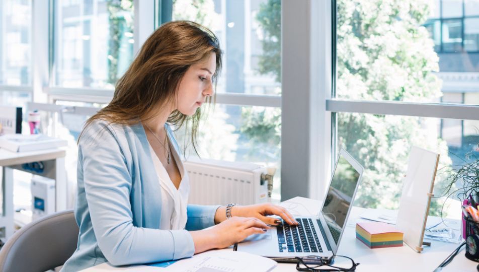 Woman in light blue blazer typing on laptop at desk by window.