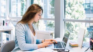 Woman working on a laptop at a desk near a window, typing.