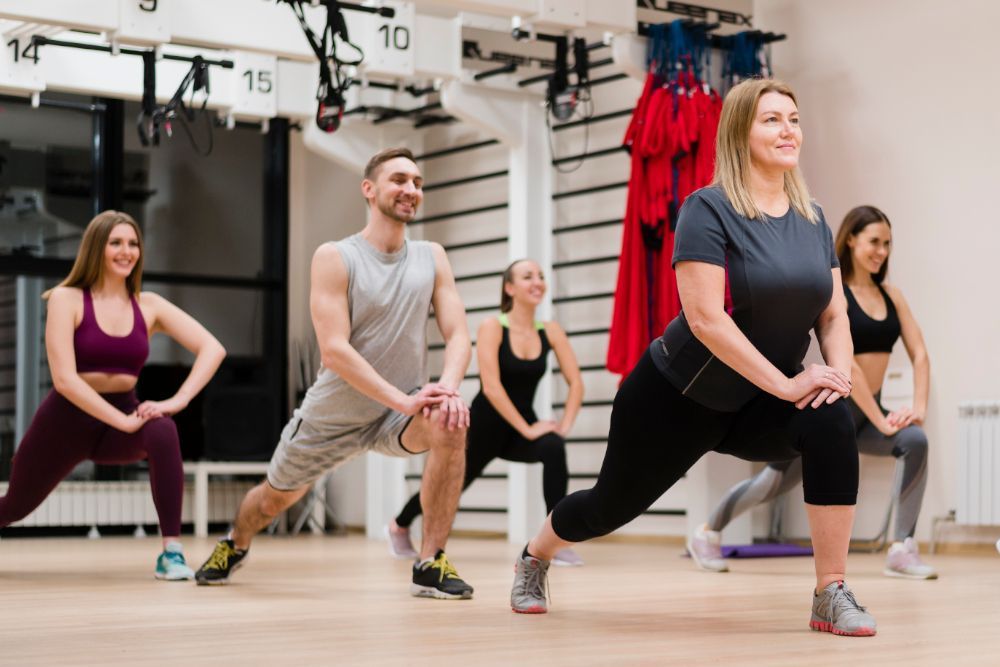 Group of five people doing lunges in a gym; they are smiling and wearing workout clothes.