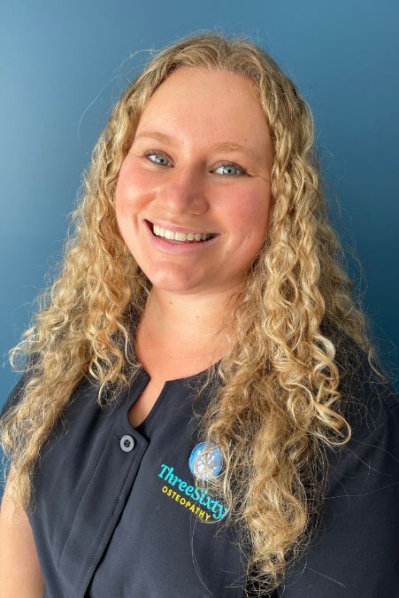 Woman with curly blonde hair smiles at the camera. She wears a navy shirt with a logo, standing in front of a teal wall.