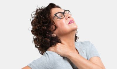 Woman with curly hair wearing glasses, clutching her neck and shoulder in pain, eyes closed.