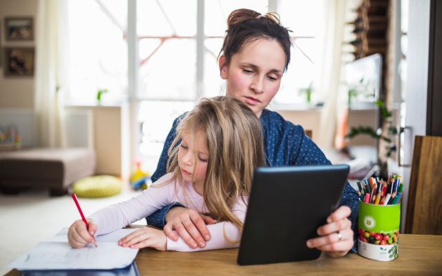 Woman and child sit at table. Child writes, while woman holds tablet, both focused, at home.